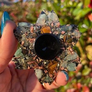 May include: A flower-shaped orgone energy generator with a black center and a mix of green, gold, and clear crystals. Copper wire is woven throughout the design. The object is held in a hand with blue nail polish.