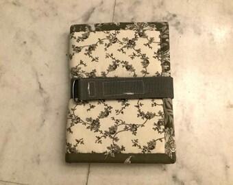 Puede incluir: Una funda de diario de tela acolchada con un estampado floral en tonos grises y crema. La funda se asegura con una correa gris y una hebilla metálica. Los bordes están ribeteados con una tela gris más oscura.