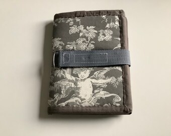 Puede incluir: Un diario de tela gris y blanca con un estampado toile de Jouy, con querubines y motivos florales. Tiene un cierre de correa gris y está bordeado con una tela gris más oscura. El diseño es clásico.