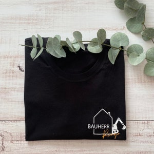 Könnte beinhalten: Schwarzes T-Shirt mit einem weißen Logo eines Hauses und eines Baggers mit dem Text "Bauherr" und einer goldenen Unterschrift.