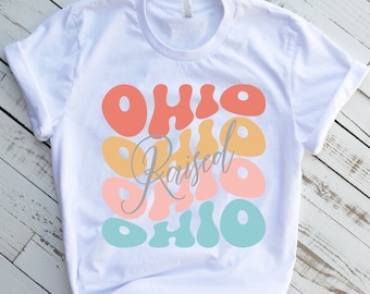 Könnte beinhalten: Weißes T-Shirt mit einem Grafikdesign des Wortes "Ohio", das in verschiedenen Farben und Größen wiederholt wird. Der Text "Raised" ist in grauer Kursivschrift zwischen den Wörtern "Ohio" geschrieben.