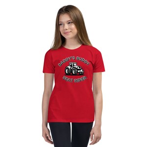 May include: Red youth t-shirt with a black and white graphic of a tractor. The graphic says "Daddy's Buddy Seat Rider".