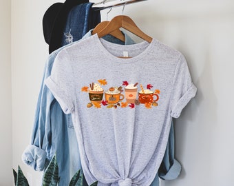 Puede incluir: Camiseta gris claro con un diseño otoñal de cuatro tazas de café con crema batida, palitos de canela y hojas de otoño. Las tazas son naranjas, marrones y rojas. El texto "Pumpkin Spice" está en una taza.