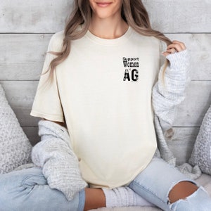 May include: A cream colored t-shirt with the text "Support Women in AG" printed on the front.