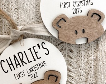 May include: Two round white wooden ornaments featuring a brown wooden bear face cutout at the bottom. One ornament is inscribed with BAKER'S FIRST CHRISTMAS 2025 and the other with CHARLIE'S FIRST CHRISTMAS 2022 in black text. Each has a jute twine loop.