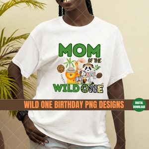 K&ouml;nnte beinhalten: Wei&szlig;es T-Shirt mit einer gr&uuml;nen und braunen Grafik, auf der "Mom of the Wild One" steht, mit einem L&ouml;wen, Zebra, Giraffe, Panda und Elefant. Die Grafik ist von Palmen und Ranken umgeben.