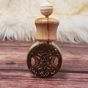May include: A clear glass bottle with a wooden stopper and a metal triple moon and pentacle design. The bottle is on a wooden surface.