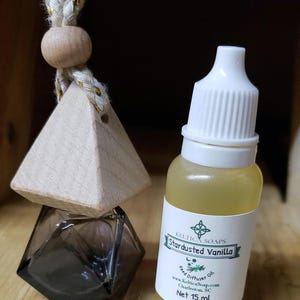 May include: A small glass bottle with a white cap filled with a clear liquid. The bottle has a white label with the text "Keltica Soaps Star dusted Vanilla Reed Diffuser Oil www.kelticasoap.com Charleston, SC Net 15 ml". The bottle is sitting on a wooden surface next to a wooden triangle with a hole in the top and a rope attached.