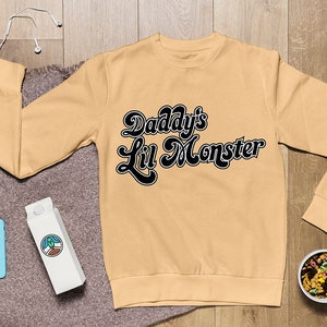 May include: A tan sweatshirt with black text that reads "Daddy's Lil Monster" in a retro font.