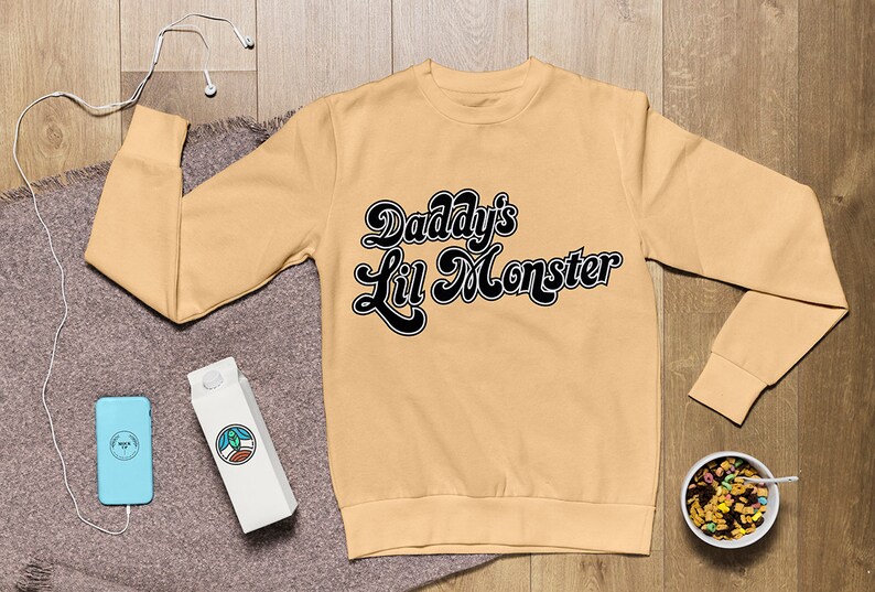 May include: A tan sweatshirt with black text that reads "Daddy's Lil Monster" in a retro font.