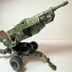 May include: A green toy artillery cannon with black wheels and a pivoting base. The cannon has a long barrel and is angled upwards. The words "SENSOR ACCESS" and "CAUTION" are visible on the side. The words "UNITED" are also visible.