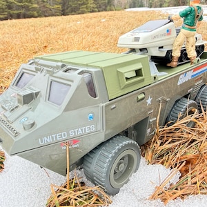 May include: A vintage olive green toy military vehicle with the text "UNITED STATES" and "G.I. JOE". A small white snowmobile with the American flag is on top, and a toy soldier is standing on the snowmobile.