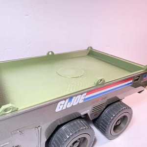 May include: A green and gray toy truck with the words "G.I. Joe" printed on the side. The truck has large, gray wheels and a flatbed with a circular indentation. Red, white, and blue stripes are visible on the side.