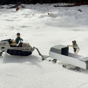 Pode incluir: Uma mota de neve de brinquedo vintage com uma figura em miniatura a conduzi-la, puxando um trenó a condizer. A mota de neve é branca com esteiras pretas e a inscrição "UNITED STATES" na lateral. O trenó também é branco e cinzento.