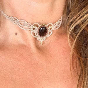 May include: Cream-colored macrame choker necklace with a dark brown oval gemstone center. The necklace features intricate knotting and small gold-colored beads. A small pendant hangs below the gemstone.