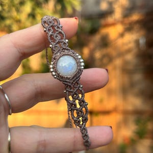 Puede incluir: Pulsera de macramé hecha a mano con una piedra lunar redonda e iridiscente en el centro. La pulsera está hecha con cordón marrón y adornada con pequeñas cuentas plateadas alrededor de la piedra lunar. El diseño de macramé es visible.