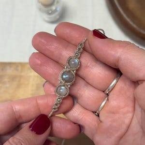 May include: A handmade macrame bracelet featuring three round, iridescent moonstone beads. The bracelet is crafted with a beige cord and small silver-colored beads. The design is simple and elegant, suitable for everyday wear. The bracelet is held in a hand.