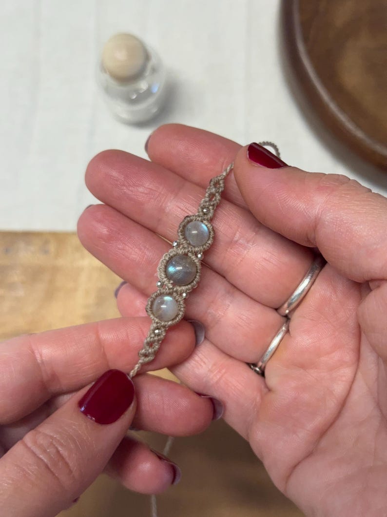 May include: A handmade macrame bracelet featuring three round, iridescent moonstone beads. The bracelet is crafted with a beige cord and small silver-colored beads. The design is simple and elegant, suitable for everyday wear. The bracelet is held in a hand.