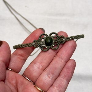 Puede incluir: Collar de macram&eacute; verde oliva con una piedra central verde oscuro. El collar presenta intrincados nudos y peque&ntilde;as cuentas de color plateado. El collar se muestra sobre un fondo claro.
