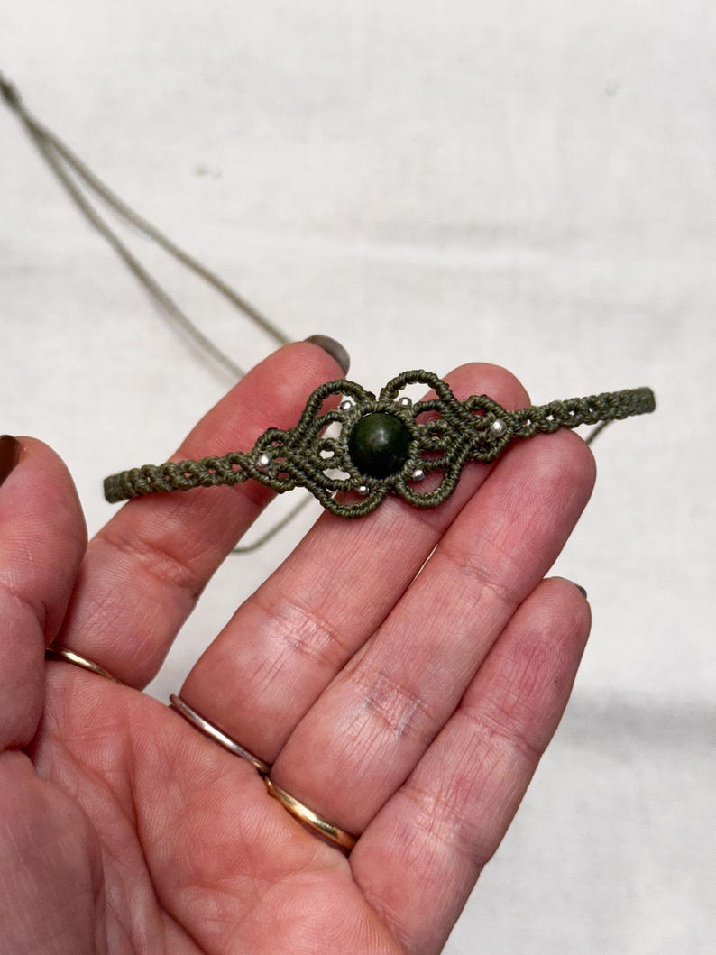 Puede incluir: Collar de macram&eacute; verde oliva con una piedra central verde oscuro. El collar presenta intrincados nudos y peque&ntilde;as cuentas de color plateado. El collar se muestra sobre un fondo claro.