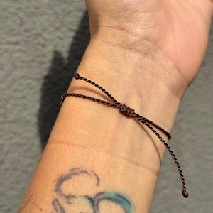 May include: A dark brown adjustable bracelet tied on a wrist. The bracelet is made of thin, woven material. The wrist has a tattoo of a symbol in shades of purple and blue. The background is a solid gray.