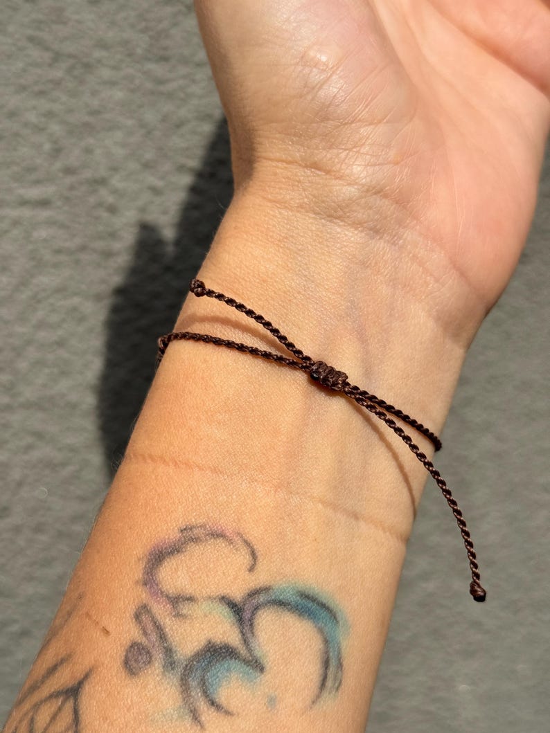 May include: A dark brown adjustable bracelet tied on a wrist. The bracelet is made of thin, woven material. The wrist has a tattoo of a symbol in shades of purple and blue. The background is a solid gray.