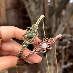 Puede incluir: Se muestran dos pulseras de macram&eacute;, una verde oliva y otra rosa empolvado. Cada pulsera presenta una piedra preciosa central, una verde y otra blanca, rodeada de intrincados nudos y peque&ntilde;as cuentas doradas. Las pulseras se sostienen sobre un fondo borroso al aire libre.