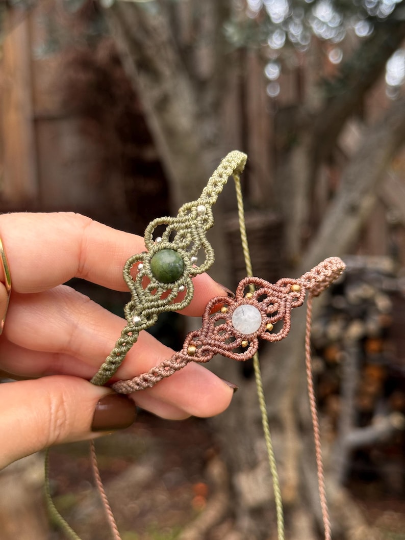 Puede incluir: Se muestran dos pulseras de macram&eacute;, una verde oliva y otra rosa empolvado. Cada pulsera presenta una piedra preciosa central, una verde y otra blanca, rodeada de intrincados nudos y peque&ntilde;as cuentas doradas. Las pulseras se sostienen sobre un fondo borroso al aire libre.