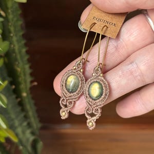 Puede incluir: Un par de pendientes hechos a mano con piedras preciosas ovaladas engastadas en macramé. Los pendientes son de color marrón claro con ganchos dorados. La etiqueta dice "EQUINOX".