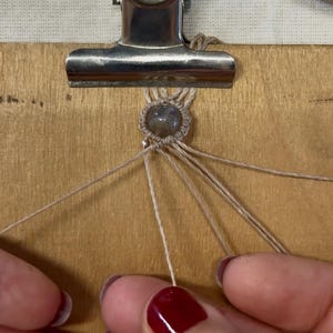 May include: Close-up of a macrame project in progress. A small, round, gray gemstone is centered within a woven pattern of beige cord. The cord is held in place by a silver clip on a wooden surface. Hands with red nail polish are visible.