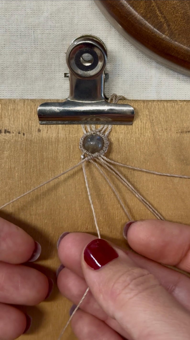 May include: Close-up of a macrame project in progress. A small, round, gray gemstone is centered within a woven pattern of beige cord. The cord is held in place by a silver clip on a wooden surface. Hands with red nail polish are visible.