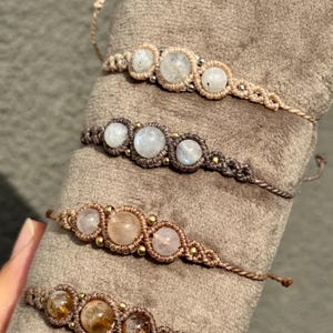 May include: Close-up of several macrame bracelets. Each bracelet features three round, light-colored gemstones, such as moonstone or quartz, set within a woven, neutral-toned macrame design. The bracelets are displayed on a soft, brown surface.