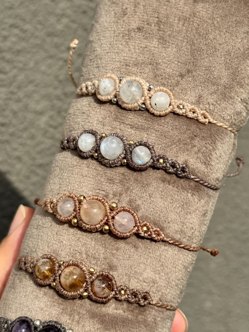 May include: Close-up of several macrame bracelets. Each bracelet features three round, light-colored gemstones, such as moonstone or quartz, set within a woven, neutral-toned macrame design. The bracelets are displayed on a soft, brown surface.