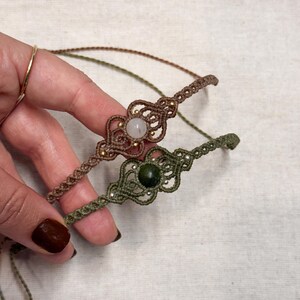 Puede incluir: Dos pulseras de macram&eacute;. Una es marr&oacute;n con una piedra clara y cuentas doradas, la otra es verde oliva con una piedra verde oscuro y cuentas plateadas. Las pulseras est&aacute;n hechas a mano y tienen intrincados patrones de nudos.