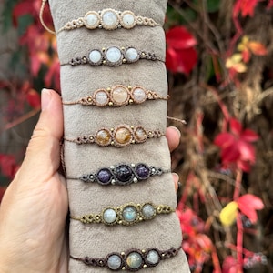 Könnte beinhalten: Eine Sammlung handgefertigter Makramee-Armbänder, jedes mit drei Edelsteinperlen. Die Armbänder sind in verschiedenen Farben erhältlich, darunter Beige, Braun und Gold, mit Edelsteinen in Weiß, Lila und Bernstein. Die Armbänder werden auf einer grauen Oberfläche präsentiert.