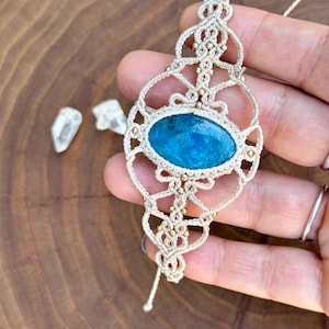 May include: A macrame pendant featuring an oval, vibrant blue stone. The pendant is crafted with beige cord and accented with small gold-colored beads. The design has an open, lacy pattern, and a cord extends from the top for hanging.