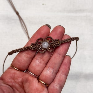 Puede incluir: Un collar gargantilla de macram&eacute; hecho a mano en tonos marrones, con un dise&ntilde;o central con una piedra de color claro y peque&ntilde;as cuentas doradas. El collar se muestra sobre un fondo claro.