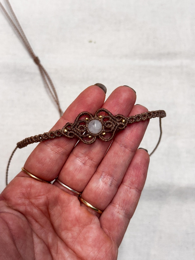 Puede incluir: Un collar gargantilla de macram&eacute; hecho a mano en tonos marrones, con un dise&ntilde;o central con una piedra de color claro y peque&ntilde;as cuentas doradas. El collar se muestra sobre un fondo claro.