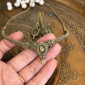 Puede incluir: Un collar de macramé y un par de pendientes a juego hechos con cordón verde oliva, pequeñas cuentas doradas y piedras de labradorita ovaladas. El collar tiene un colgante central con un adorno dorado en forma de lágrima en la parte inferior.