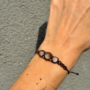 May include: A brown macrame bracelet featuring three clear, round gemstones. The bracelet is adorned with small, gold-colored beads and is displayed on a person's arm.