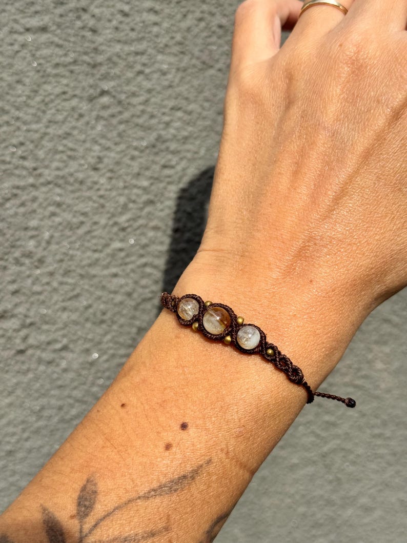May include: A brown macrame bracelet featuring three clear, round gemstones. The bracelet is adorned with small, gold-colored beads and is displayed on a person's arm.
