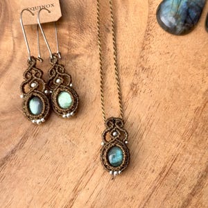 K&ouml;nnte beinhalten: Eine braune Schnurkette mit einem Anh&auml;nger aus gr&uuml;nem Labradorit. Der Anh&auml;nger ist von einem braunen Makramee-Muster umgeben. Die Kette liegt auf einer Holzoberfl&auml;che mit zwei passenden Ohrringen mit gr&uuml;nen Labradorit-Steinen.
