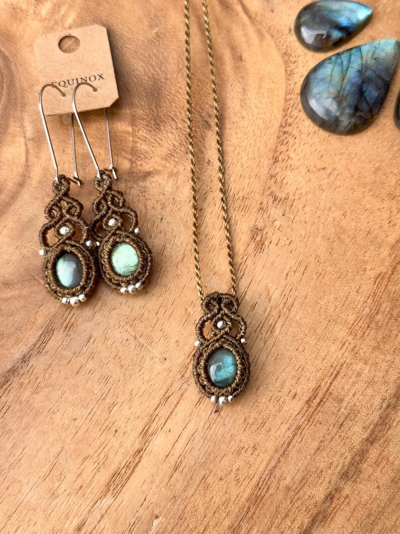 K&ouml;nnte beinhalten: Eine braune Schnurkette mit einem Anh&auml;nger aus gr&uuml;nem Labradorit. Der Anh&auml;nger ist von einem braunen Makramee-Muster umgeben. Die Kette liegt auf einer Holzoberfl&auml;che mit zwei passenden Ohrringen mit gr&uuml;nen Labradorit-Steinen.