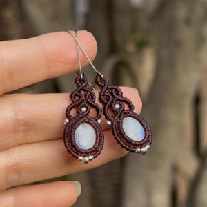 Puede incluir: Un par de pendientes de macramé hechos a mano. Los pendientes presentan un diseño tejido marrón oscuro con una piedra central ovalada de color azul claro. Se incorporan pequeñas cuentas plateadas y los pendientes tienen ganchos plateados.