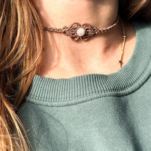 Puede incluir: Un collar gargantilla de macram&eacute; marr&oacute;n con una piedra blanca central y detalles dorados. El intrincado dise&ntilde;o tejido presenta un patr&oacute;n sim&eacute;trico. El collar se usa sobre una parte superior verde, con cabello largo y casta&ntilde;o claro visible.