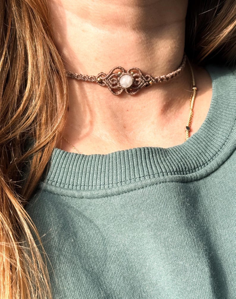 Puede incluir: Un collar gargantilla de macram&eacute; marr&oacute;n con una piedra blanca central y detalles dorados. El intrincado dise&ntilde;o tejido presenta un patr&oacute;n sim&eacute;trico. El collar se usa sobre una parte superior verde, con cabello largo y casta&ntilde;o claro visible.