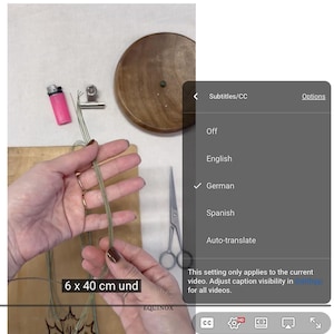 May include: A person's hands holding green twine, with a pink lighter, scissors, and a wooden disc visible. The text "6 x 40 cm und" is displayed on the screen. The video settings show German subtitles.
