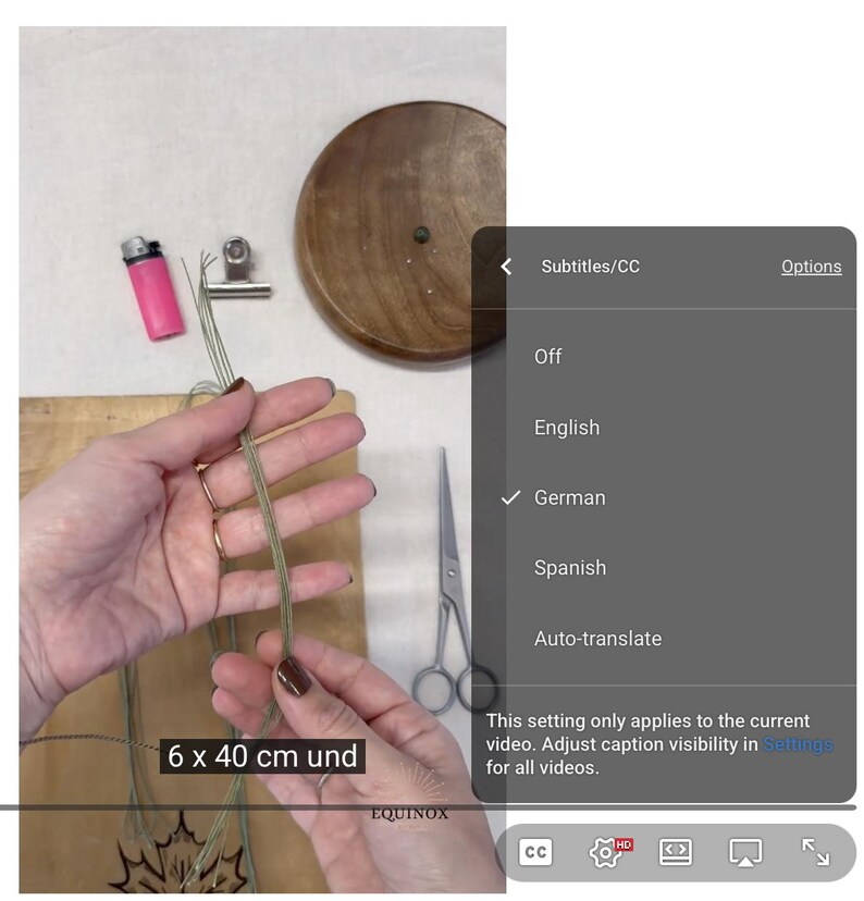 May include: A person's hands holding green twine, with a pink lighter, scissors, and a wooden disc visible. The text "6 x 40 cm und" is displayed on the screen. The video settings show German subtitles.