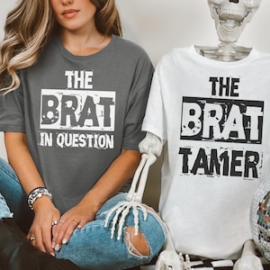 Könnte beinhalten: Zwei T-Shirts, eines dunkelgrau und eines weiß, mit dem Text "THE BRAT IN QUESTION" und "THE BRAT TAMER" in einer kräftigen, verwitterten Schriftart. Die Shirts werden an einer Person und einem Skelett gezeigt.