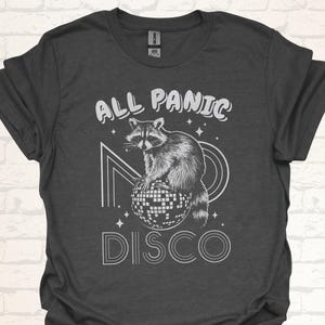 Op de afbeelding: Een donkergrijs T-shirt met een witte afbeelding van een wasbeer die op een discobal zit. De tekst "All Panic Disco" is in wit boven de wasbeer gedrukt.